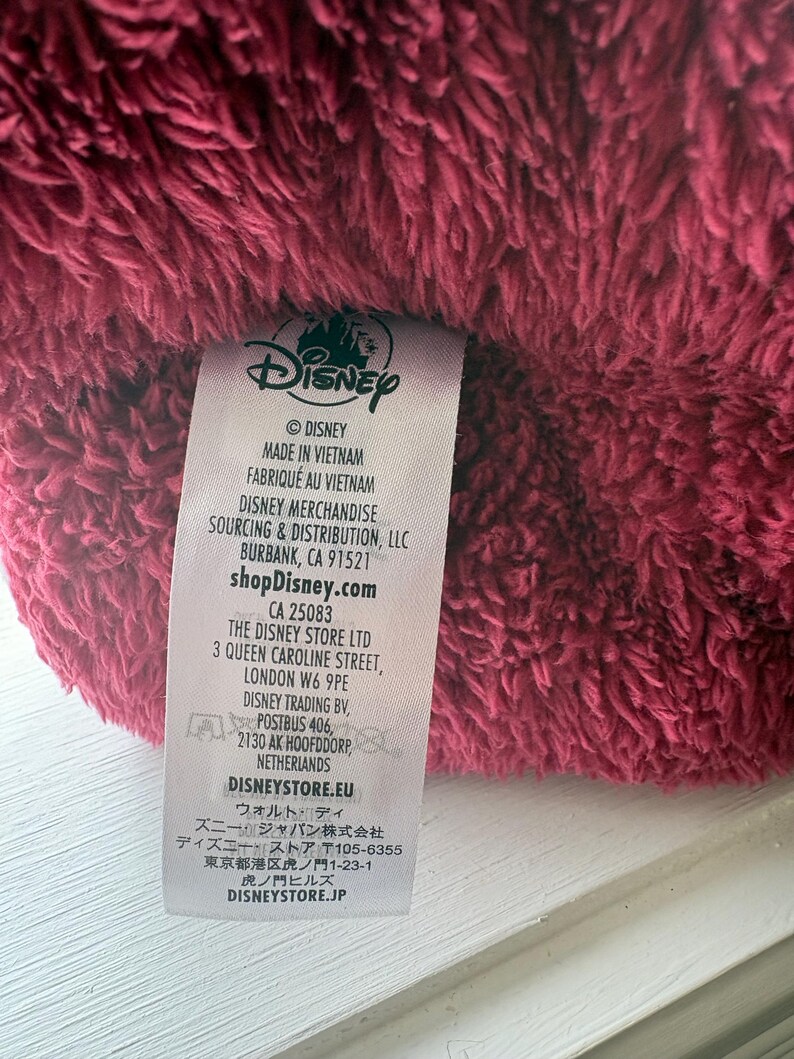 May include: Close-up of a plush, burgundy-colored fabric with a Disney label attached. The label includes the Disney logo, manufacturing details, and website information. The fabric appears soft and textured.