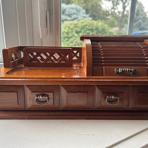 May include: A vintage wooden desk organizer with a roll-top compartment and three drawers. The organizer has a dark brown finish, silver-toned drawer pulls, and a decorative lattice design on the top. It is a classic desk accessory.