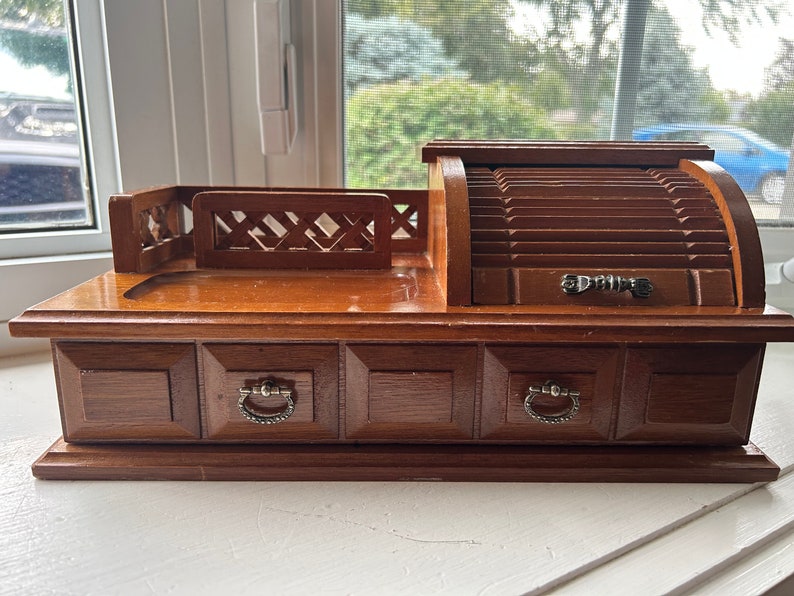 May include: A vintage wooden desk organizer with a roll-top compartment and three drawers. The organizer has a dark brown finish, silver-toned drawer pulls, and a decorative lattice design on the top. It is a classic desk accessory.