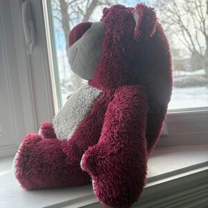 May include: A plush teddy bear in shades of burgundy, white, and pink. The bear is sitting on a windowsill, with its side profile visible. The bear has a soft, fuzzy texture and a friendly expression.
