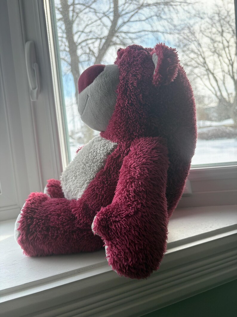 May include: A plush teddy bear in shades of burgundy, white, and pink. The bear is sitting on a windowsill, with its side profile visible. The bear has a soft, fuzzy texture and a friendly expression.