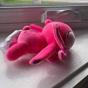 May include: A bright pink plush bunny toy with long floppy ears and purple accents on its paws. The stuffed animal is lying on a white windowsill, with a tag attached.
