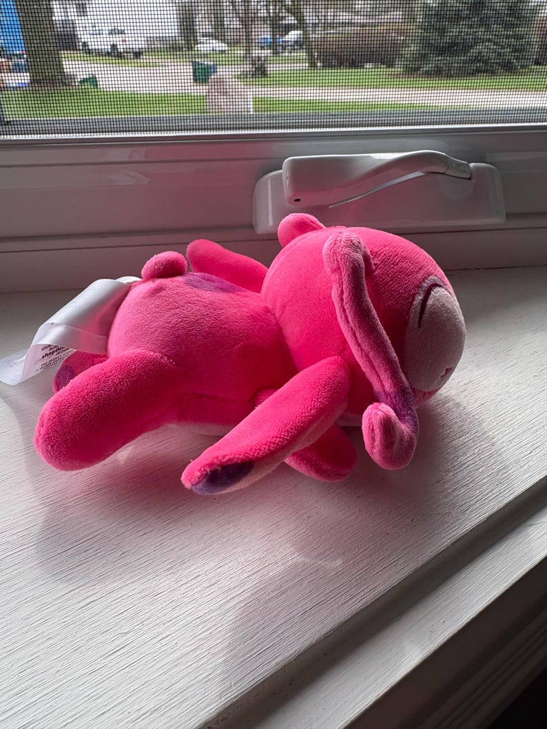 May include: A bright pink plush bunny toy with long floppy ears and purple accents on its paws. The stuffed animal is lying on a white windowsill, with a tag attached.