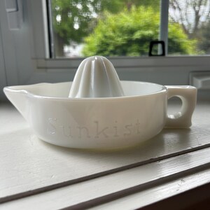 Peut inclure: Presse-agrumes vintage en verre blanc avec un presse-agrumes et un bec verseur. Le mot "Sunkist" est en relief sur le côté. Le presse-agrumes est posé sur une surface blanche près d'une fenêtre.