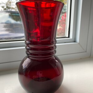 Peut inclure: Un vase en verre rouge rubis avec une base arrondie et un col évasé. Le col présente des rainures horizontales. Le verre transparent laisse passer la lumière. Le vase est placé près d'une fenêtre.