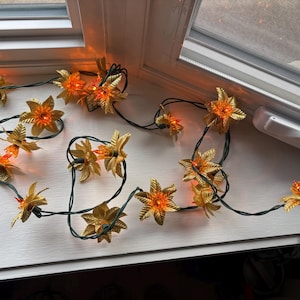 May include: A string of decorative lights featuring gold-coloured flower-shaped ornaments with orange-coloured bulbs. The lights are strung on a dark green wire and are arranged on a white windowsill. The lights are turned on.