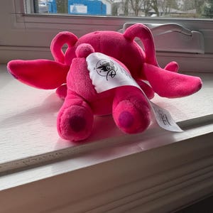 May include: A bright pink plush toy, possibly a character from a movie or cartoon, with large ears and a tag. The toy is lying on a white surface near a window. The tag has a logo and text.