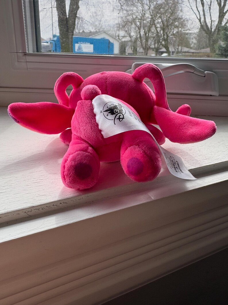 May include: A bright pink plush toy, possibly a character from a movie or cartoon, with large ears and a tag. The toy is lying on a white surface near a window. The tag has a logo and text.