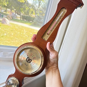 May include: A wall-mounted weather station featuring a dark wood frame with a brass-rimmed barometer, a thermometer with a red liquid indicator, and a small hygrometer at the base. The barometer dial displays the Taylor brand name and pressure readings.