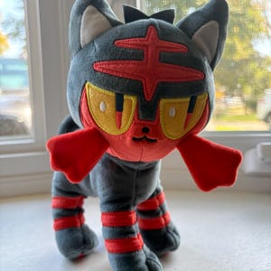 May include: A plush toy of a cat-like creature with dark gray fur, a red face mask, and yellow eyes. It features red markings on its forehead, red cheek tufts, and two red stripes on each of its four legs. The plush has pointed ears with white inner sections and a small black nose.