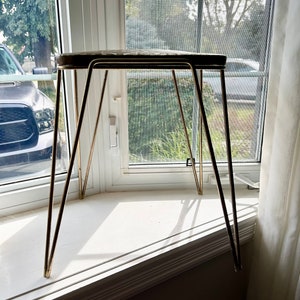 May include: A gold metal stool with a round, brown top and three legs. The stool is sitting on a white windowsill.