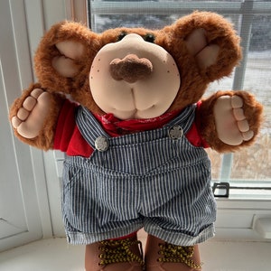 Puede incluir: Un oso de peluche marrón que lleva un mono de trabajo a rayas azules y blancas, una camisa roja y botas marrones. El oso tiene la nariz marrón y los ojos verdes.