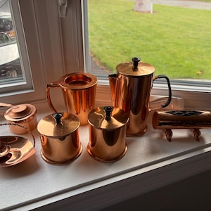 May include: A collection of copper kitchenware, including a coffee pot, sugar bowls, and small saucepans. The items are arranged on a windowsill, reflecting natural light. The copper has a warm, reddish-brown hue.