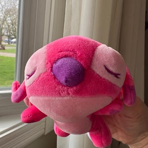 May include: A plush toy in shades of pink and purple, featuring a rounded body, small arms and legs, and large, closed eyes. The toy has a textured, soft appearance and is held in a person's hand.