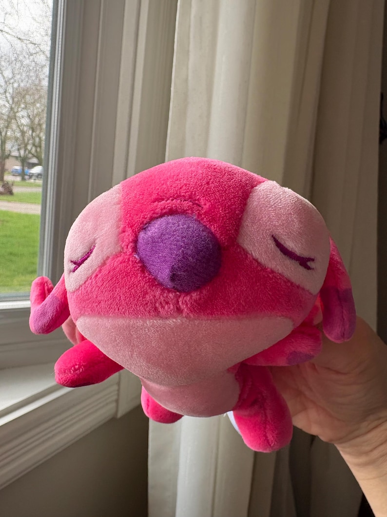May include: A plush toy in shades of pink and purple, featuring a rounded body, small arms and legs, and large, closed eyes. The toy has a textured, soft appearance and is held in a person's hand.