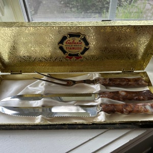 May include: A vintage carving set presented in a gold-coloured box. The set includes a carving fork and two knives with stainless steel blades and marbled brown handles. The box has a gold floral pattern and the text "Galaxie Gold Medal Stainless".