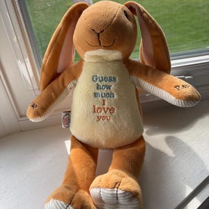 May include: A plush bunny toy in shades of brown and cream, with long ears and a soft, cuddly appearance. The bunny has the text "Guess how much I love you" printed on its chest. A charming and huggable stuffed animal.