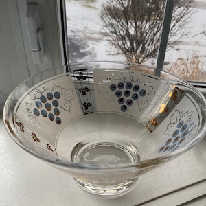May include: A clear glass bowl with a gold and white design featuring grapes and vines. The bowl has a wide rim and a flared base.