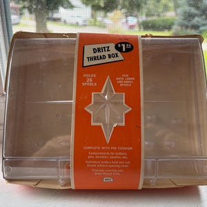 May include: A clear plastic Dritz thread box with a bright orange paper band wrapped around the center. The band features the text Dritz Thread Box, holds 26 spools, fits both large and small spools, and complete with pin cushion, with a star cutout.