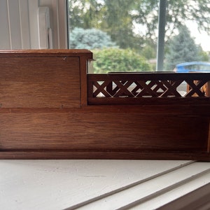 May include: A small, wooden decorative box with a dark brown finish. The box has a rectangular shape with a small compartment on the left and a decorative railing on the right. The box is sitting on a white windowsill.