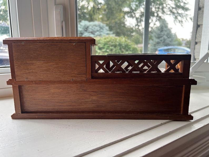 May include: A small, wooden decorative box with a dark brown finish. The box has a rectangular shape with a small compartment on the left and a decorative railing on the right. The box is sitting on a white windowsill.