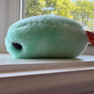 May include: A soft, mint green plush toy shaped like a microbe with a single dark eye. A red fabric tag is attached to the side, featuring the white text GIANTmicrobes, FLU, and giantmicrobes.com, indicating the product branding and identity.