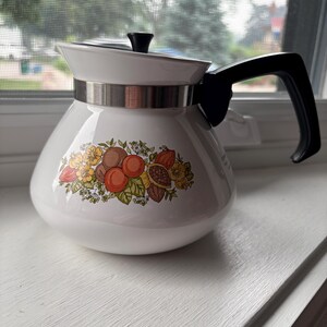May include: A white teapot with a black handle and a black lid knob. The body features a horizontal metal band and a colourful botanical illustration of various fruits, including oranges, brown nuts, and green leaves, centred on the front surface.