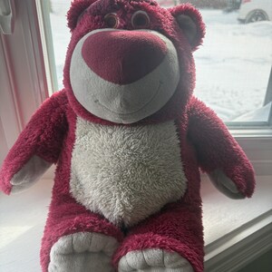 May include: A plush, pink teddy bear with a white belly and a heart-shaped nose. The bear has a friendly expression and is sitting upright. Its fur is a deep pink, and its paws and ears are a lighter shade of gray.