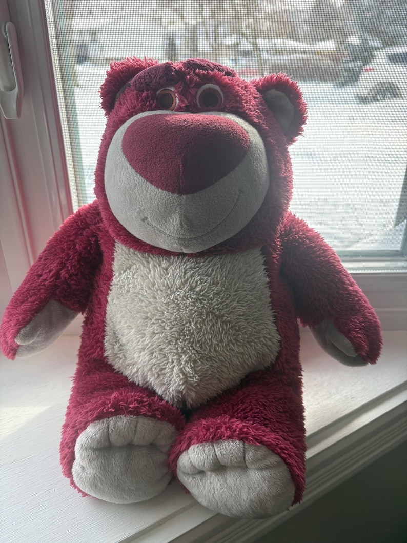 May include: A plush, pink teddy bear with a white belly and a heart-shaped nose. The bear has a friendly expression and is sitting upright. Its fur is a deep pink, and its paws and ears are a lighter shade of gray.