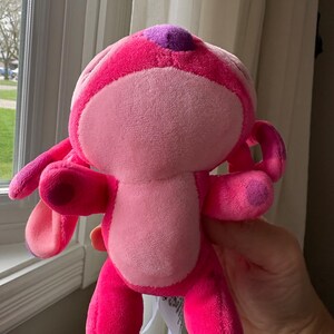 May include: A plush toy in shades of pink, featuring a rounded body, large ears, and small arms and legs. The toy has a lighter pink face and belly, with darker pink accents and a purple spot on its head. A tag is attached.