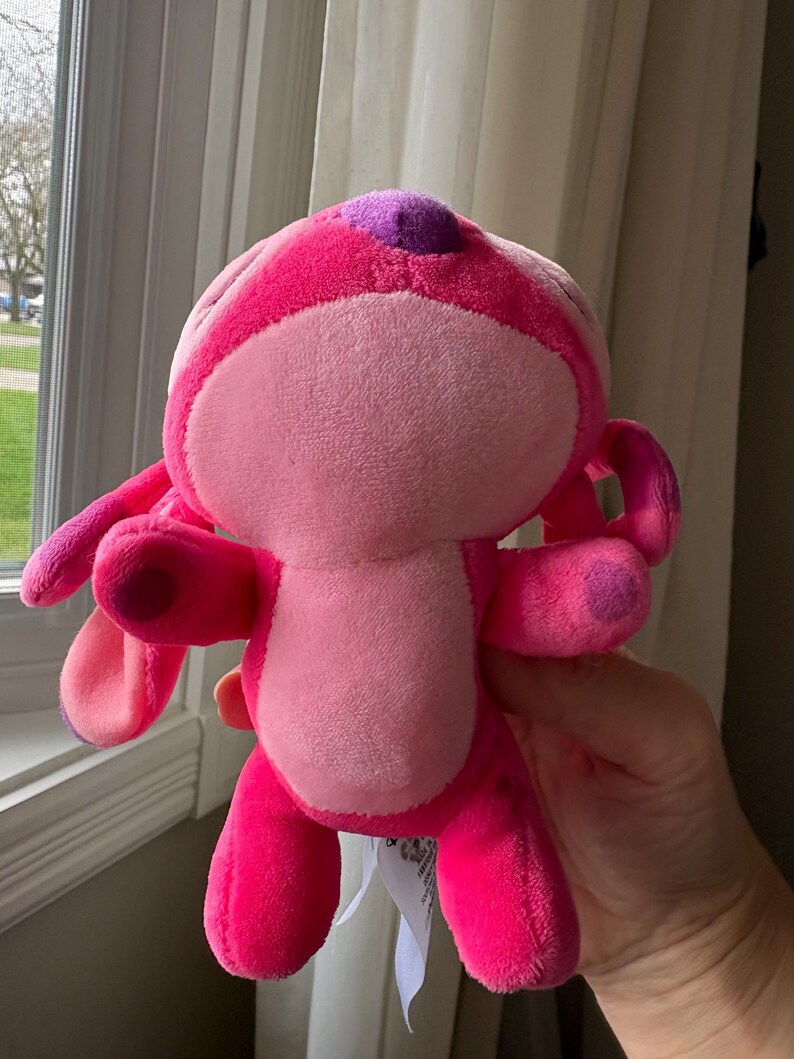 May include: A plush toy in shades of pink, featuring a rounded body, large ears, and small arms and legs. The toy has a lighter pink face and belly, with darker pink accents and a purple spot on its head. A tag is attached.