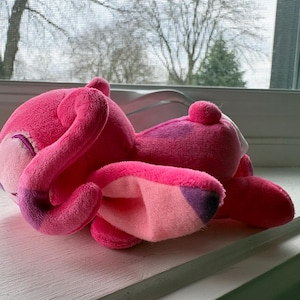May include: A plush pink flamingo toy with a curved neck and a small, round head. The flamingo has a light pink beak and a darker pink body with purple accents. The toy is lying on a white windowsill.