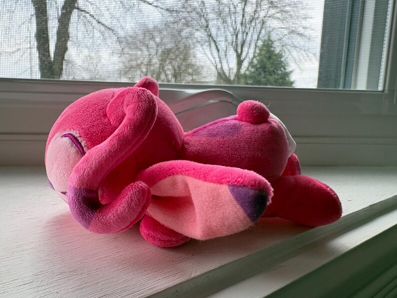 May include: A plush pink flamingo toy with a curved neck and a small, round head. The flamingo has a light pink beak and a darker pink body with purple accents. The toy is lying on a white windowsill.
