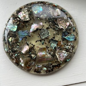 May include: Round, clear resin coaster with iridescent abalone shell pieces embedded in the resin. The coaster has a speckled, brown, and gold appearance.