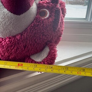 May include: A plush, pink bear with a gray belly and a brown and white eye. The bear is approximately 16 inches long, measured with a yellow tape measure. The bear is sitting on a white windowsill.
