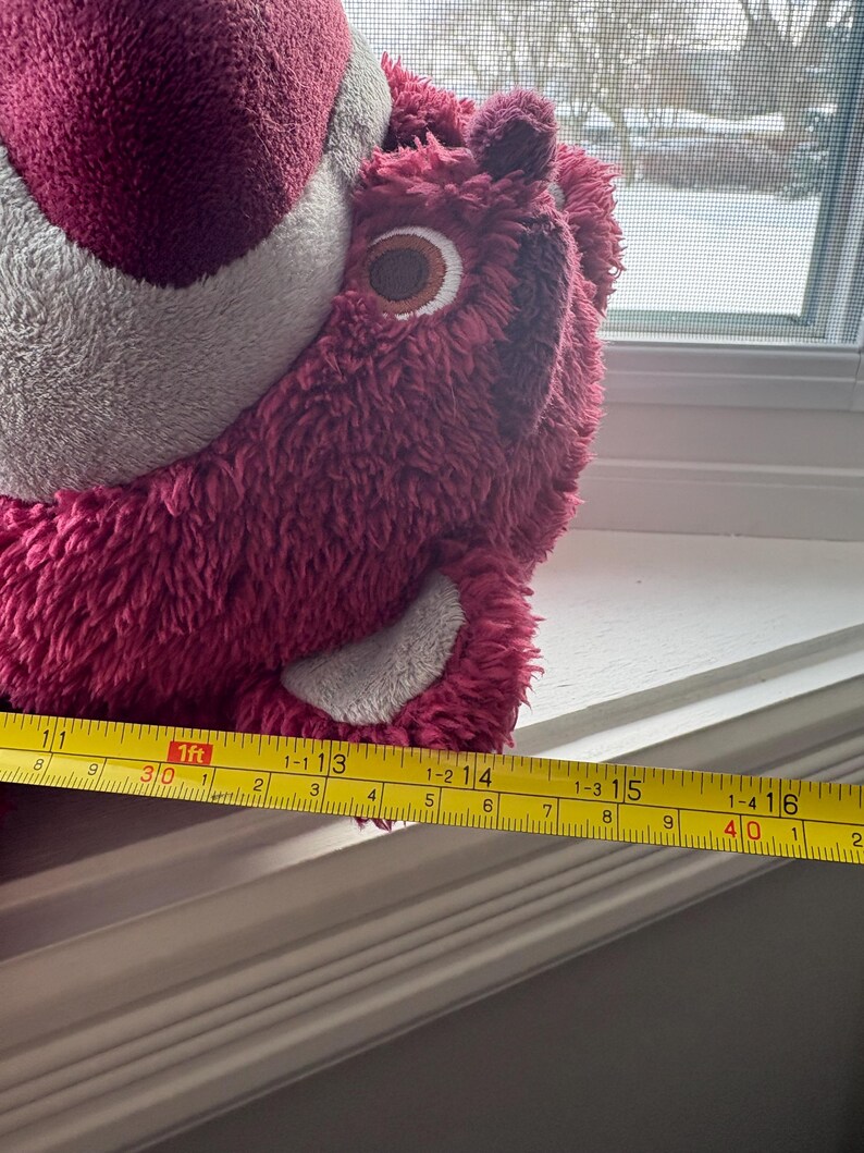 May include: A plush, pink bear with a gray belly and a brown and white eye. The bear is approximately 16 inches long, measured with a yellow tape measure. The bear is sitting on a white windowsill.