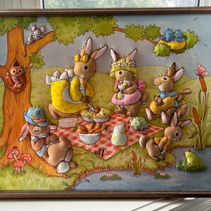 May include: A framed felt appliqu&eacute; artwork depicting a group of five bunnies having a picnic in a forest setting. The bunnies are wearing clothes and enjoying a pie, fruit, and other picnic treats. The artwork is surrounded by a brown wooden frame.