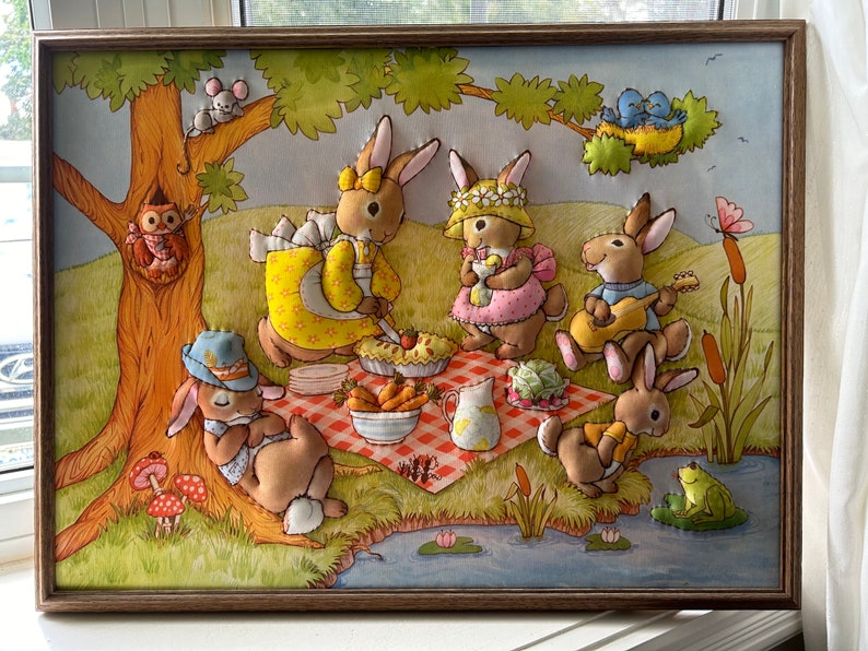 May include: A framed felt appliqu&eacute; artwork depicting a group of five bunnies having a picnic in a forest setting. The bunnies are wearing clothes and enjoying a pie, fruit, and other picnic treats. The artwork is surrounded by a brown wooden frame.