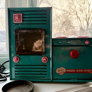 May include: Vintage teal toy oven with a matching stove top. The oven has a small window and red and pink accents. The stove top has red burners and a red knob. The oven has the text "Junior Chef MAGIC-COOL OVEN".