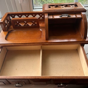May include: A wooden jewelry box with a warm brown finish. It features a top compartment with a small drawer and a larger drawer below. The top has a decorative cutout design, and the drawers have metal pulls.