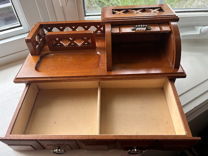 May include: A wooden jewelry box with a warm brown finish. It features a top compartment with a small drawer and a larger drawer below. The top has a decorative cutout design, and the drawers have metal pulls.