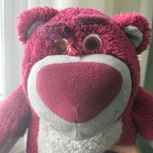 May include: A plush, pink teddy bear with a heart-shaped nose and brown eyes. The bear has a white muzzle and a slightly grumpy expression. The fur is a deep pink color, and the ears are a lighter shade of pink with gray accents.