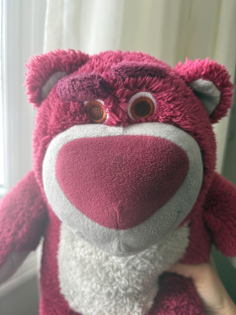 May include: A plush, pink teddy bear with a heart-shaped nose and brown eyes. The bear has a white muzzle and a slightly grumpy expression. The fur is a deep pink color, and the ears are a lighter shade of pink with gray accents.