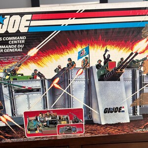 May include: A G.I. Joe Headquarters Command Center toy box featuring a graphic illustration of a military base with action figures, vehicles, and weapons. The box displays the G.I. Joe logo, red and blue stripes, and text in English and French.