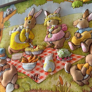 May include: A framed felt appliqu&eacute; wall art featuring five brown rabbits having a picnic. The rabbits are wearing clothes and are sitting on a red and white checkered blanket. There is a pie, a pitcher of lemonade, and a bowl of carrots on the blanket. The rabbits are surrounded by green grass and flowers.