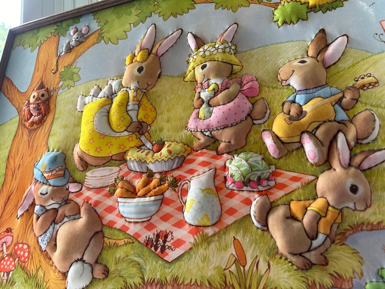 May include: A framed felt appliqu&eacute; wall art featuring five brown rabbits having a picnic. The rabbits are wearing clothes and are sitting on a red and white checkered blanket. There is a pie, a pitcher of lemonade, and a bowl of carrots on the blanket. The rabbits are surrounded by green grass and flowers.