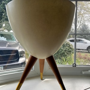 Op de afbeelding: Een witte plantenbak met een brede, ronde kom en drie houten poten met gouden puntjes. De plantenbak staat op een vensterbank.