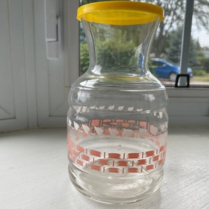 May include: A clear glass pitcher with a yellow lid and a pink and white geometric design. The pitcher is sitting on a white surface.