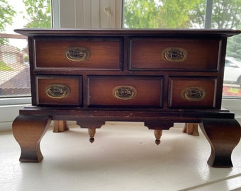 May include: A wooden jewelry box with six drawers and decorative brass pulls. The box is on four legs with a carved design.
