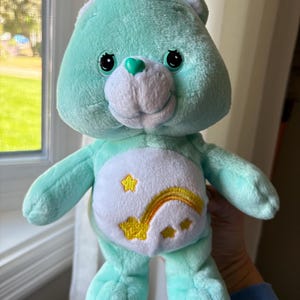 Könnte beinhalten: Ein mintgrüner Care Bear-Plüschbär mit einem weißen Bauch, der ein gelbes Sternschnuppen-Design zeigt. Der Bär hat schwarze Augen, ein grünes Herz auf der Nase und ein lächelndes Gesicht. Ein kuscheliges Spielzeug.